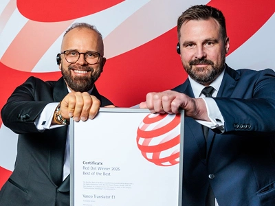 Deux hommes en tenue de travail formelle — celui de gauche en smoking et portant des lunettes, celui de droite en costume bleu foncé — tiennent ensemble, à deux mains, un certificat encadré. Le certificat porte clairement la mention « Red Dot Winner 2025 Best of the Best », décerné à « Vasco Translator E1 ». À l'arrière-plan, on aperçoit un décor graphique dynamique à grande échelle, composé de motifs abstraits ondulés rouges et blancs.