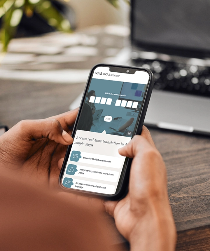 Vue par-dessus l'épaule d'une personne tenant un smartphone sur lequel s'affiche l'interface web de Vasco Audience. L'écran présente un champ de saisie intitulé « Saisissez le code de session » avec un bouton blanc « REJOINDRE », situé au-dessus d'une liste d'instructions indiquant « Accédez à la traduction en temps réel en 4 étapes simples ». En arrière-plan, on aperçoit un bureau en bois flou et un ordinateur portable.  