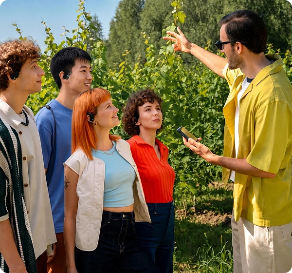Un groupe de cinq personnes se trouve en plein air dans un vignoble baigné de soleil. À droite, un homme vêtu d'une chemise jaune et portant des lunettes de soleil tient un appareil Translator Vasco Q1 et fait un geste en direction des vignes verdoyantes. À gauche, quatre personnes écoutent attentivement, chacune équipée d'un Vasco Translators E1 noir, illustrant ainsi la fonctionnalité de conversation de groupe en temps réel. 
