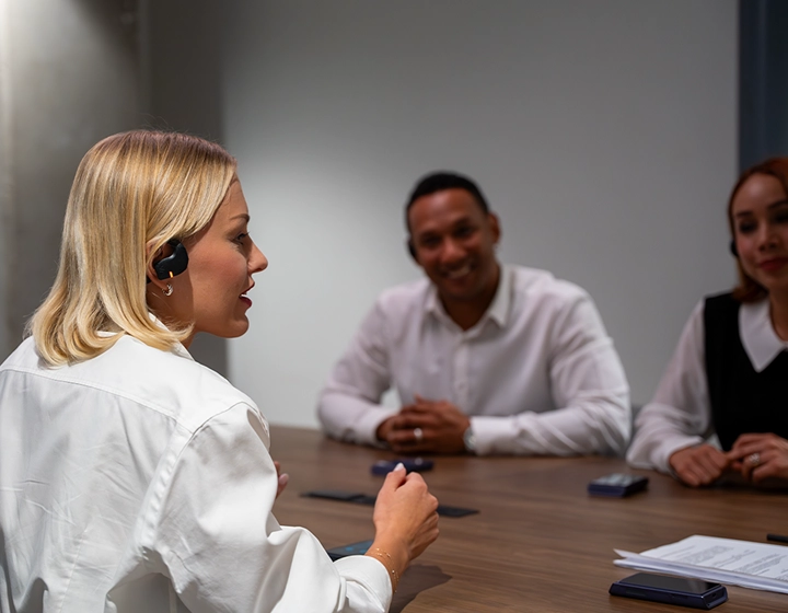 Profil d'une femme blonde vêtue d'un chemisier blanc, en train de s'exprimer lors d'une réunion d'affaires autour d'une table de conférence en bois. Elle porte un écouteur noir Vasco Translation E1. À l'arrière-plan, flou, deux autres professionnels sont assis de l'autre côté de la table ; ils sourient et écoutent attentivement. 