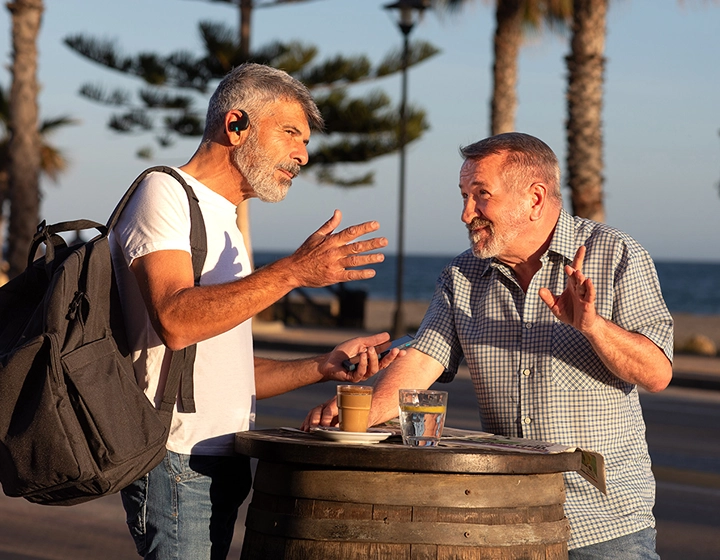 Deux hommes sont debout près d'une table en terrasse et discutent. L'un d'eux porte un sac à dos, et tous deux utilisent des écouteurs Vasco Translator E1. 