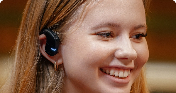 Gros plan sur le profil d'une jeune femme souriante portant un écouteur Vasco Translation E1 noir. L'image met en avant la conception hygiénique et ouverte de l'appareil, montrant qu'il repose confortablement à l'extérieur de l'oreille sans pénétrer dans le conduit auditif. 