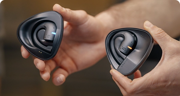 Gros plan sur des mains tenant deux étuis de recharge noirs distincts contenant des écouteurs Vasco avec fonction de traduction. L'image montre comment distinguer les deux modèles : l'un est équipé d'une LED bleue et l'autre d'une LED orange, correspondant respectivement à des locuteurs différents. 