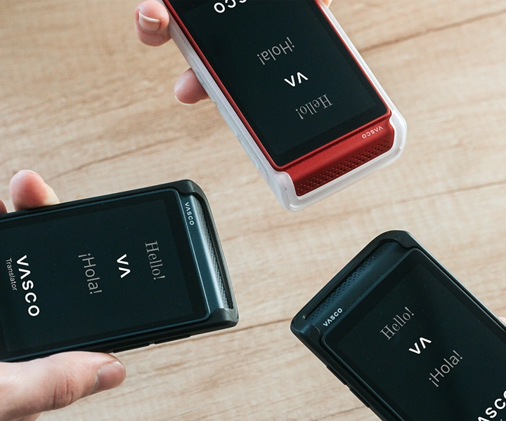 Trois personnes, tenant des appareils Vasco Translator Q1, les présentent ensemble ; les écrans affichent l’écran d’accueil avec le logo Vasco et les mots « Hello ! » et « ¡Hola ! ». Les appareils sont proposés dans des étuis en silicone disponibles en trois couleurs : Solid Black, Dark Caramel et Milky White.