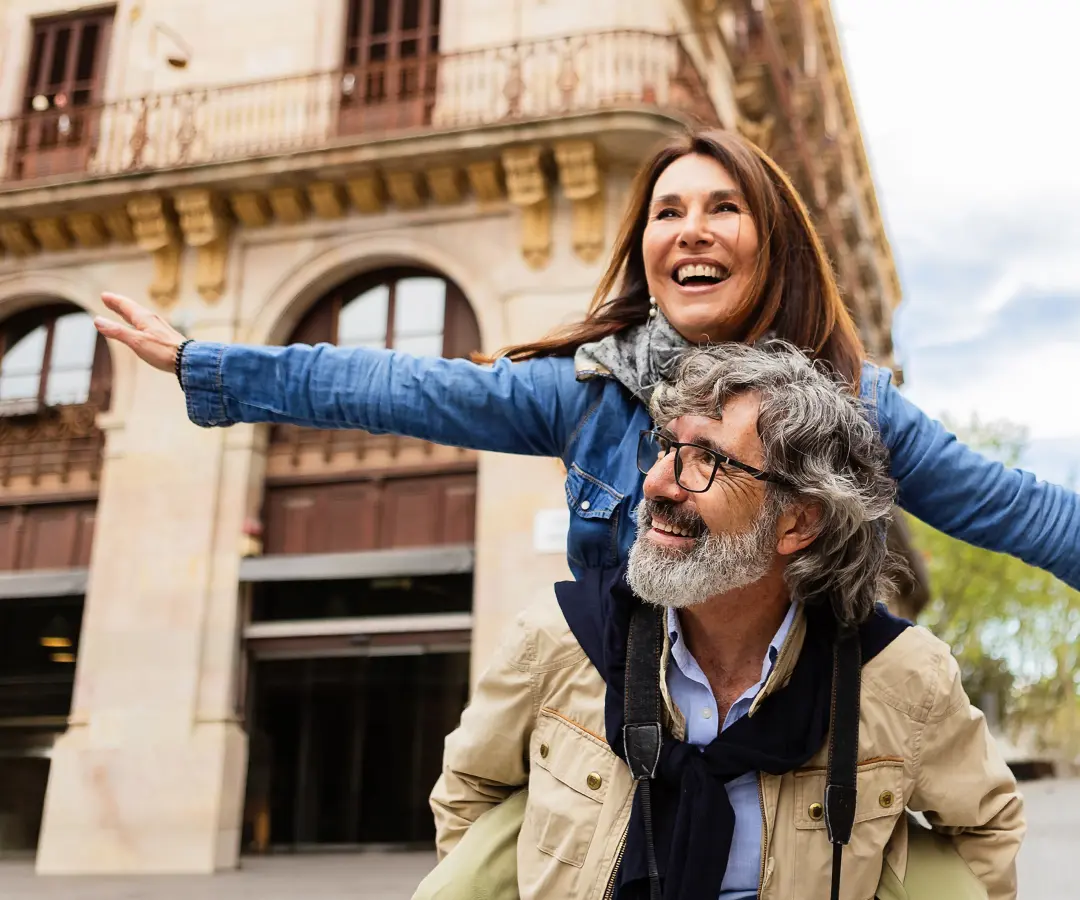 Dans une rue d'une ville européenne, un homme d'âge mûr, tout sourire, porte une femme sur son dos ; tous deux arborent un large sourire tandis qu'elle pointe du doigt au loin, le bras levé. Il porte des lunettes, une veste beige et un appareil photo, tandis qu'elle porte une veste en jean et une écharpe. 