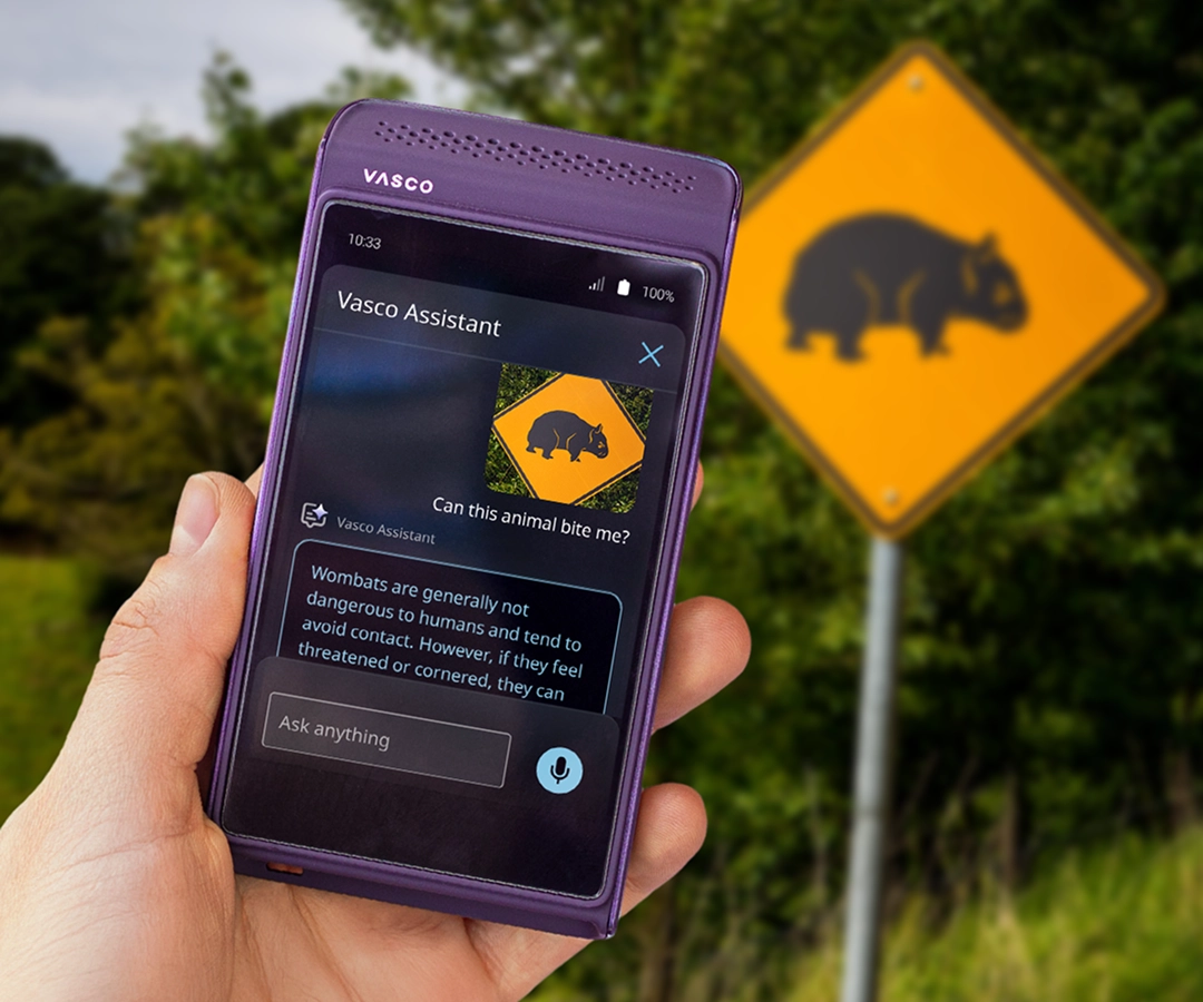 Une main tenant un traducteur électronique Vasco violet. L'application Vasco Assistant est visible sur l'écran de l'appareil, affichant une photo d'un panneau d'avertissement avec l'image d'un wombat et une description de ces animaux. À l'arrière-plan, derrière la main tenant l'appareil, se trouve une route et le même panneau routier jaune avec un wombat, qui apparaît sur l'écran du traducteur.