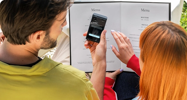 Vue par-dessus l'épaule d'un homme et d'une femme aux cheveux orange vif, assis à une table de restaurant et consultant un menu. L'homme tient un Vasco Translator Q1 noir sur lequel s'affiche un écran « Partager » comportant un champ de saisie permettant d'envoyer la traduction par e-mail. On aperçoit en arrière-plan une nappe blanche et le décor du restaurant.